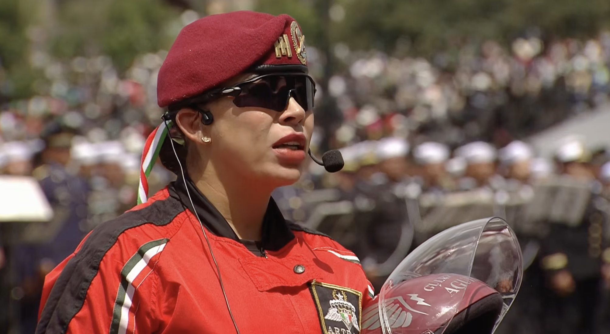Primer plano de la capitán segunda intendente paracaidista Jessica Lozano Ortega, integrante del equipo de salto libre Guerreros Azteca"de la Brigada de Fusileros Paracaidistas, rindiendo el parte de novedades a la Comandanta Suprema de las Fuerzas Armadas. Luce boina guinda, uniforme rojo de salto, micrófono con diadema y sostiene su casco de protección tras aterrizar en el Zócalo capitalino.