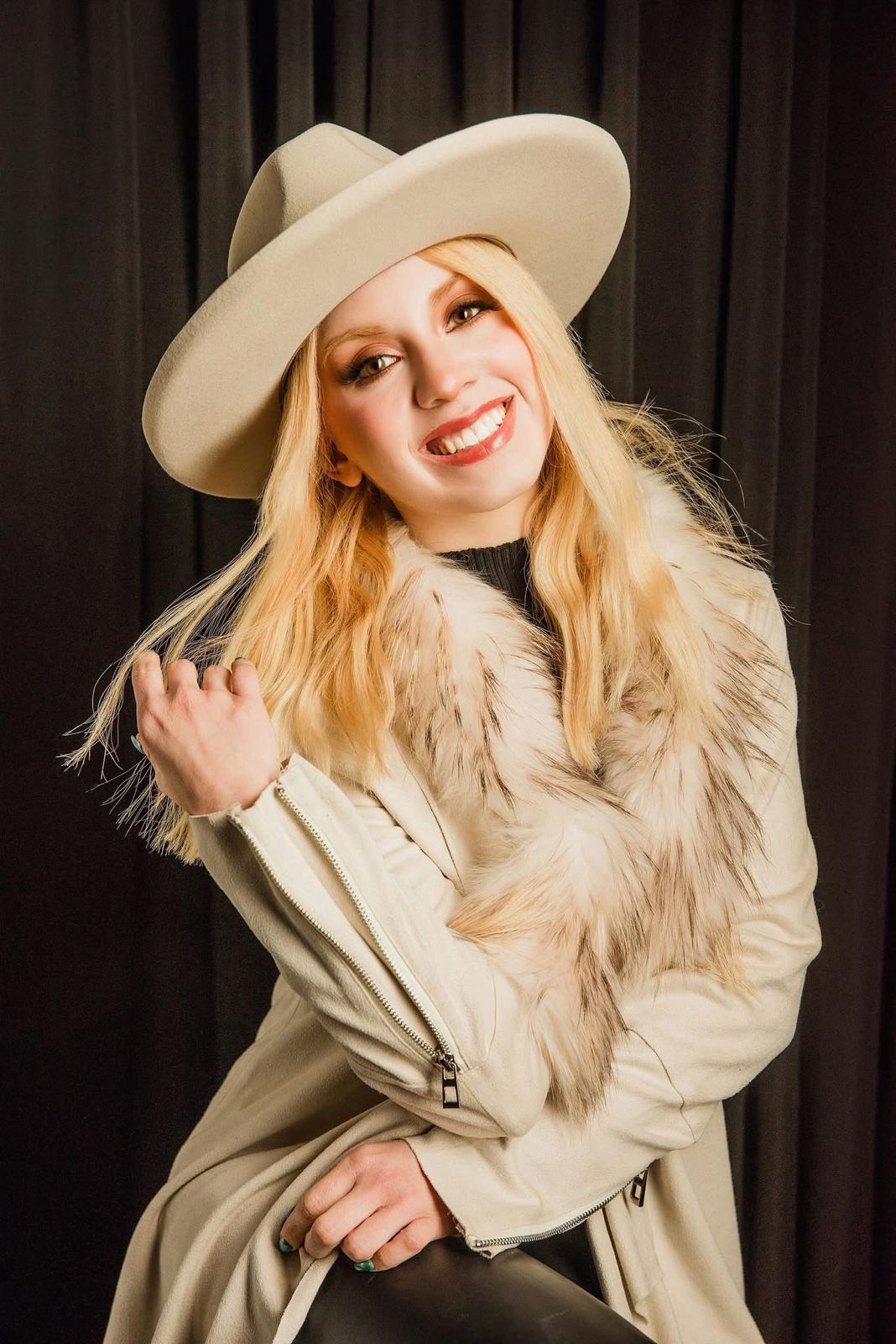 Retrato sonriente de Karla Deister, con cabello rubio, luciendo un sombrero de fieltro beige y una chaqueta de color claro con cuello de piel sintética sobre un fondo oscuro de cortinas. Ella mira a la cámara y sostiene un mechón de su cabello.