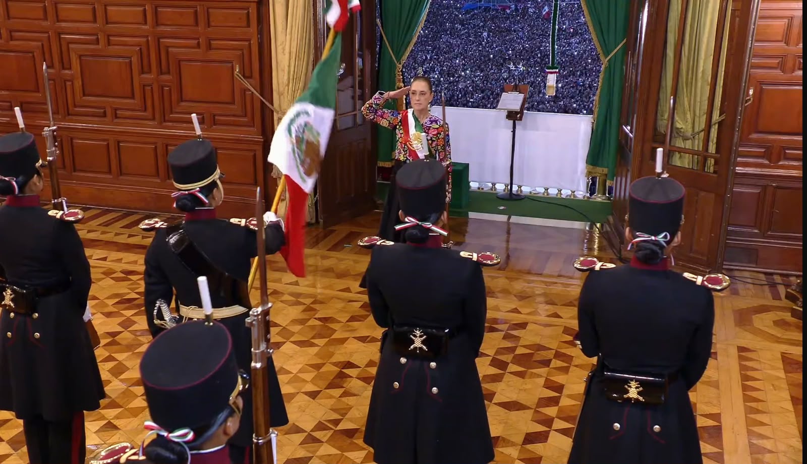 Claudia Sheinbaum realiza el saludo militar frente a la escolta del Heroico Colegio Militar al recibir la bandera de México en un salón de Palacio Nacional, previo a la ceremonia del Grito de Independencia, con la multitud visible en el Zócalo al fondo.