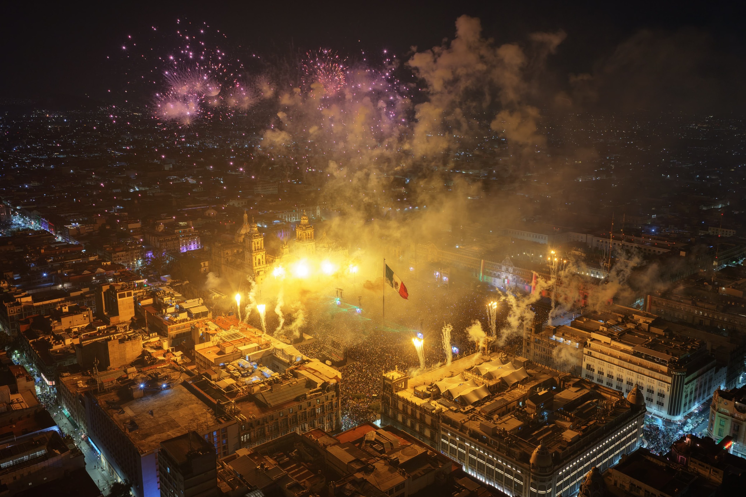 Espectacular toma nocturna de la Plaza de la Constitución de la Ciudad de México rebosante de energía, con el cielo lleno de fuegos artificiales y la bandera nacional ondeando en el centro, capturando la atmósfera festiva de la Independencia de México.