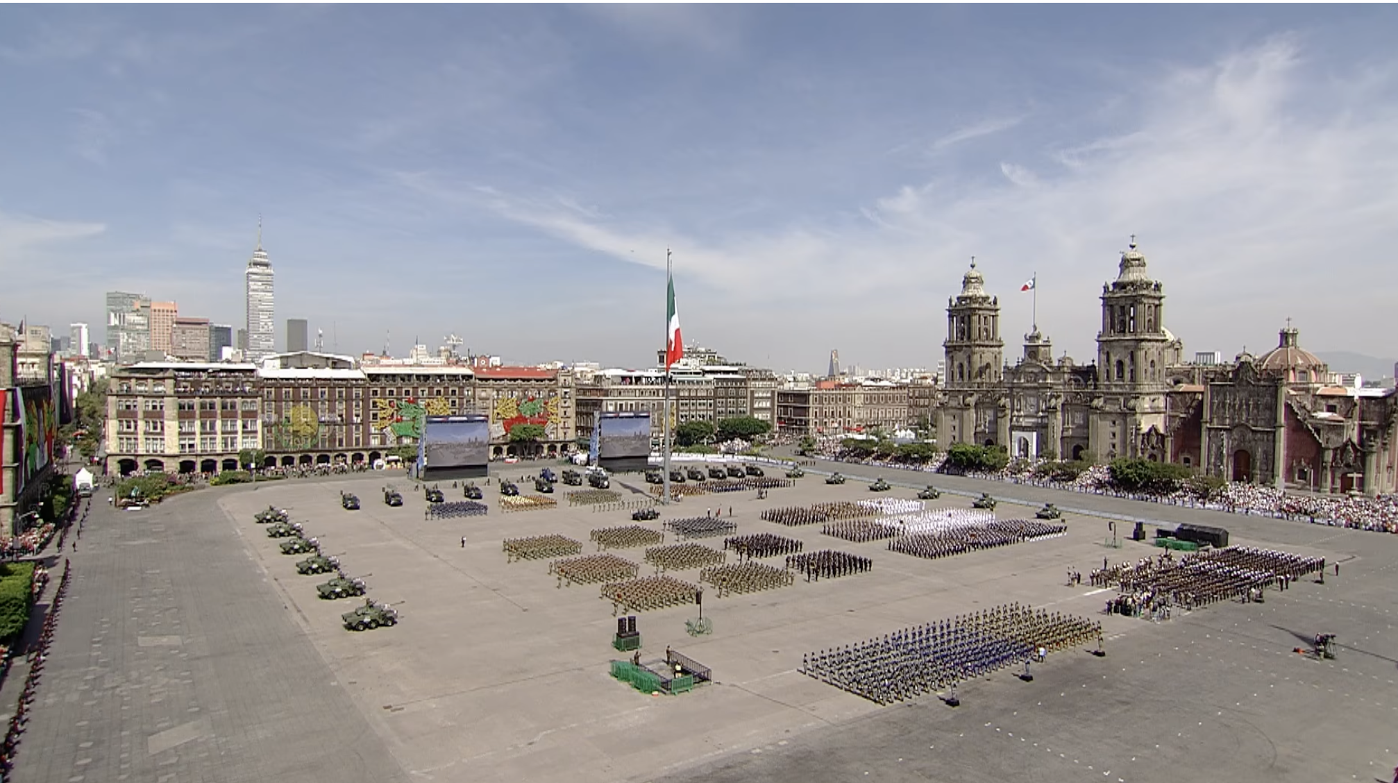Soldados mexicanos en formación militar, vistos desde atrás, en la Plaza de la Constitución (Zócalo) de Ciudad de México. Se distinguen uniformes con patrones de camuflaje azul y verde. De fondo, se aprecian el Palacio Nacional y la bandera de México junto a un asta bandera monumental