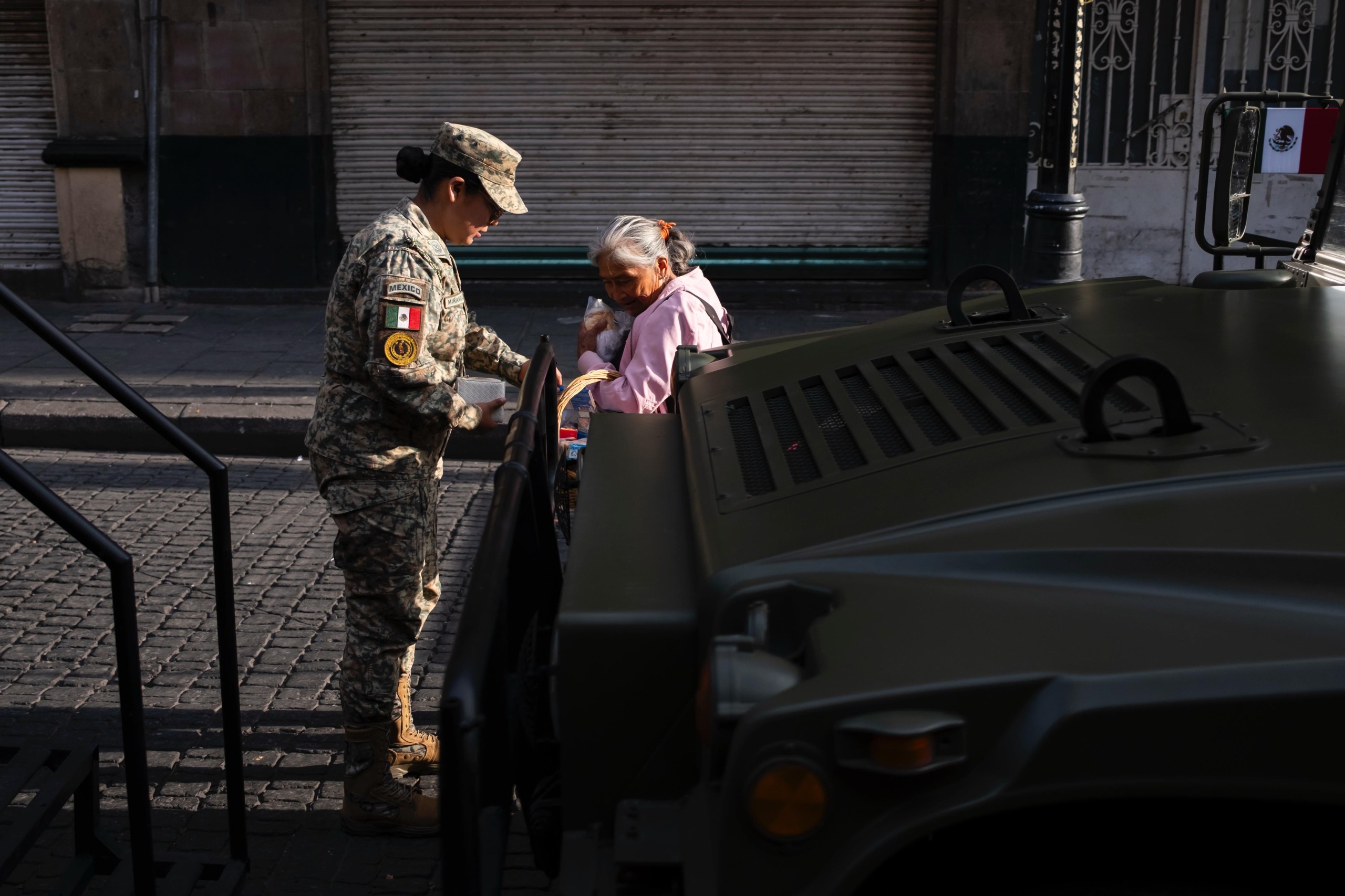 Una mujer militar del Ejército Mexicano interactúa amablemente con una mujer adulta mayor en la calle, junto a un vehículo táctico militar estacionado durante un operativo o evento público.