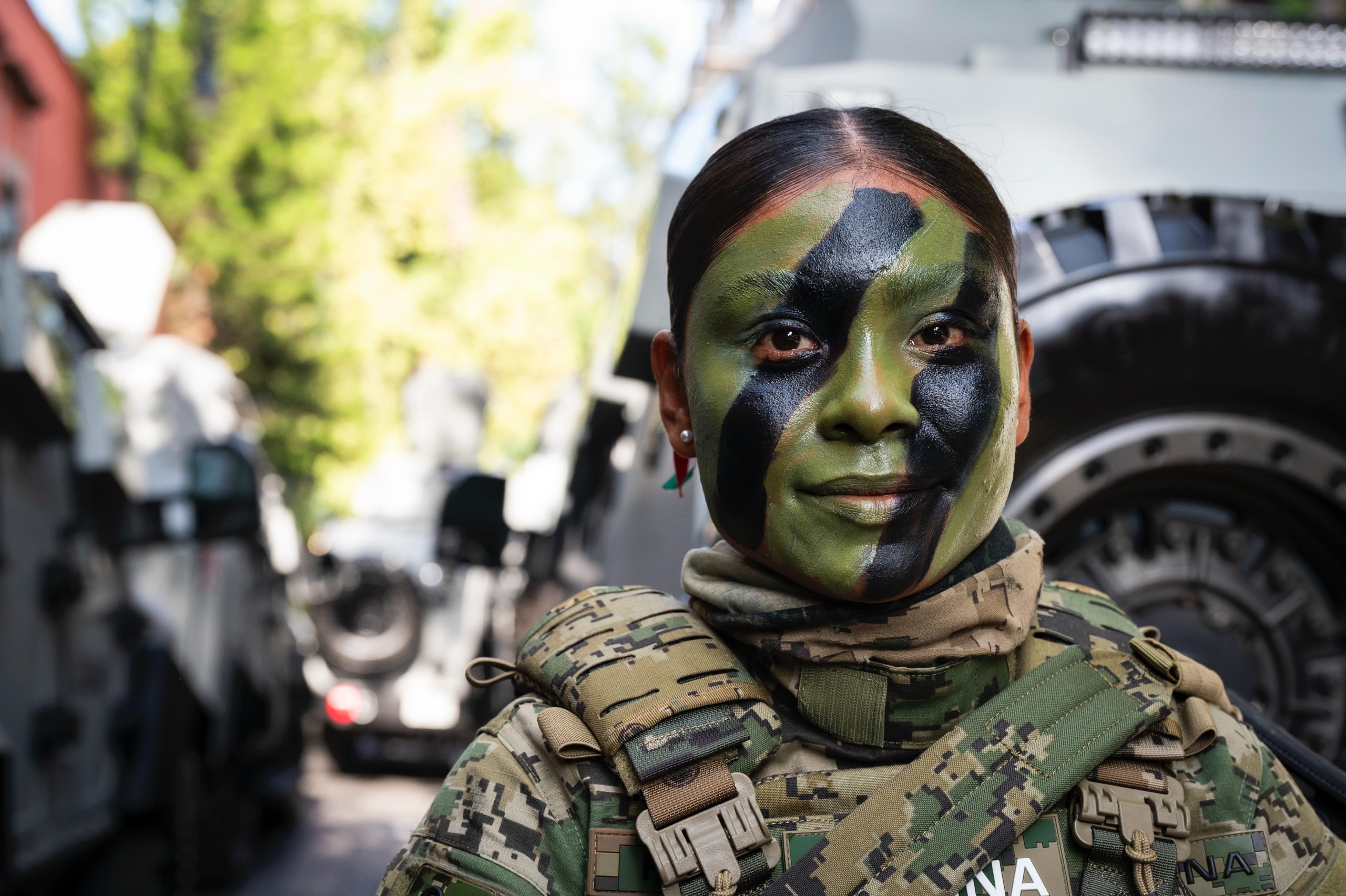 Retrato en primer plano de una mujer militar del Ejército Mexicano (SEDENA) con la cara pintada con camuflaje verde y negro, portando uniforme táctico. De fondo se aprecian vehículos blindados militares estacionados.