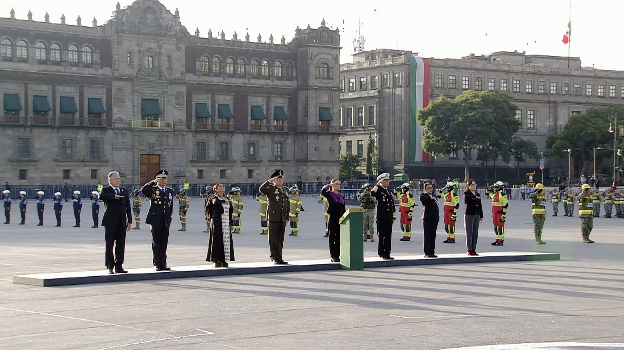 Una ceremonia oficial se lleva a cabo en la Plaza de la Constitución de la Ciudad de México (Zócalo), con el Palacio Nacional de fondo. Un grupo de líderes gubernamentales y militares mexicanos, vestidos formalmente y de uniforme, están de pie en un estrado mientras hacen el saludo a la bandera