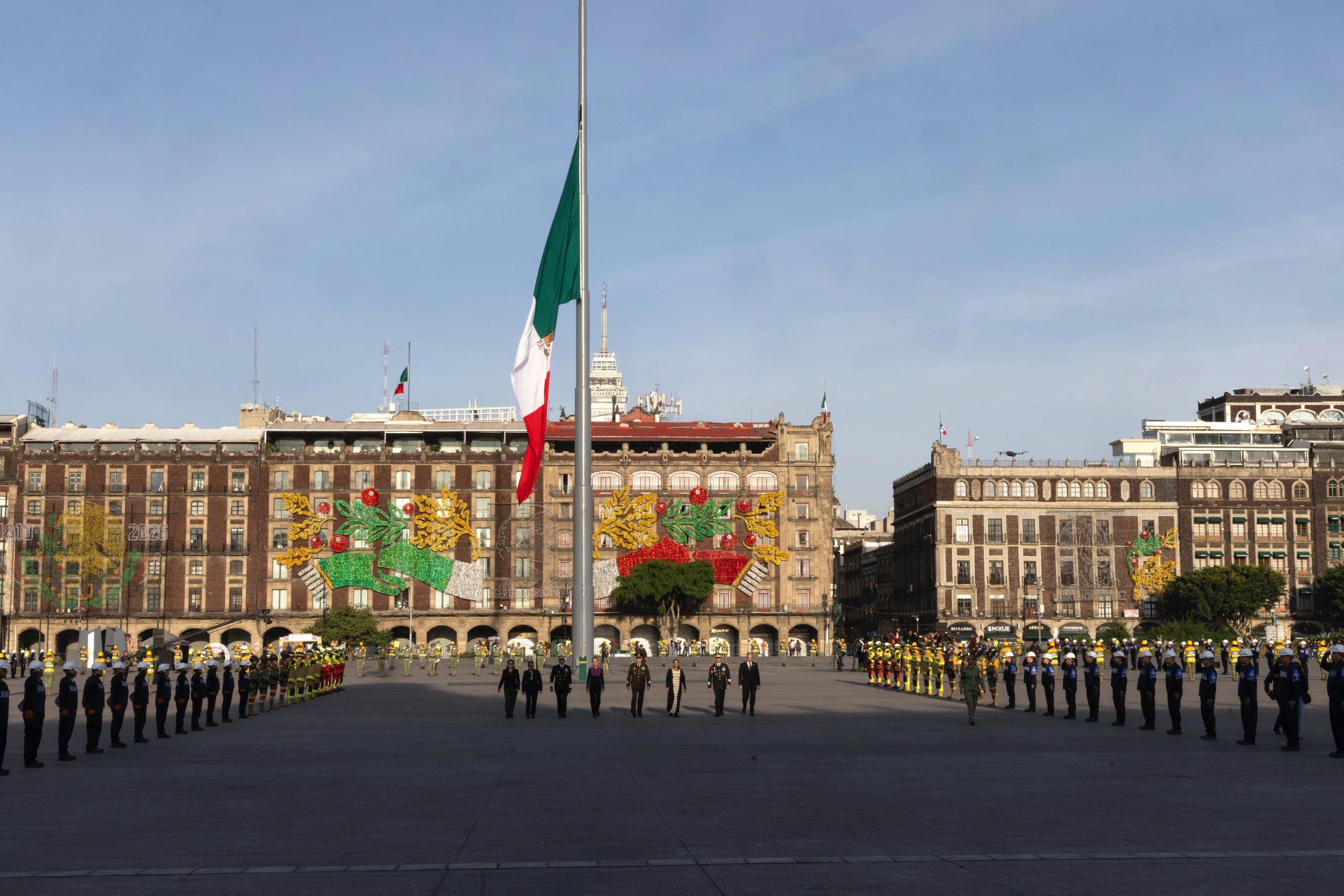 Autoridades encabezadas por la presidenta y mandos militares caminan hacia el asta bandera del Zócalo de la CDMX, con la bandera de México a media asta y contingentes de emergencia a los lados.