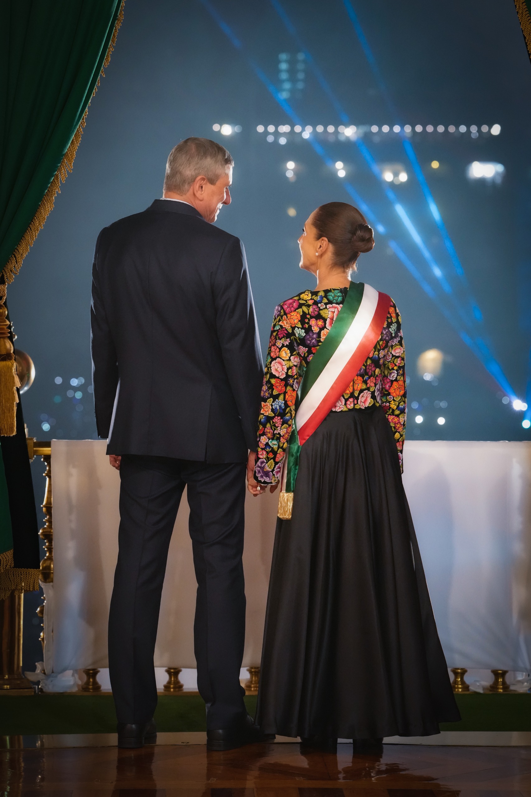 Claudia Sheinbaum y su esposo Jesús María Tarriba tomados de la mano y vistos desde atrás en el balcón de Palacio Nacional. Ella viste un vestido tradicional bordado con la banda presidencial y él porta un traje oscuro, observando la Plaza de la Constitución iluminada durante la ceremonia del Grito de Independencia.