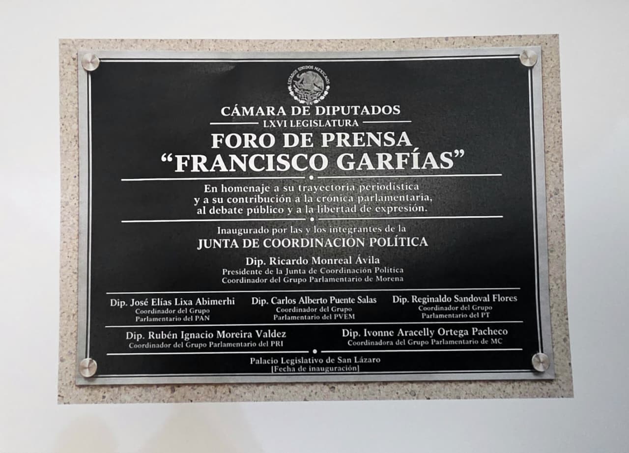Una foto de primer plano de una placa conmemorativa de metal oscuro fijada a una pared texturizada con cuatro tornillos metálicos. En la parte superior, presenta el escudo de armas de México. El texto principal en mayúsculas dice: "CÁMARA DE DIPUTADOS", "LXVI LEGISLATURA", "FORO DE PRENSA", “FRANCISCO GARFÍAS”. Debajo, el texto de homenaje: "En homenaje a su trayectoria periodística y a su contribución a la crónica parlamentaria, al debate público y a la libertad de expresión.
