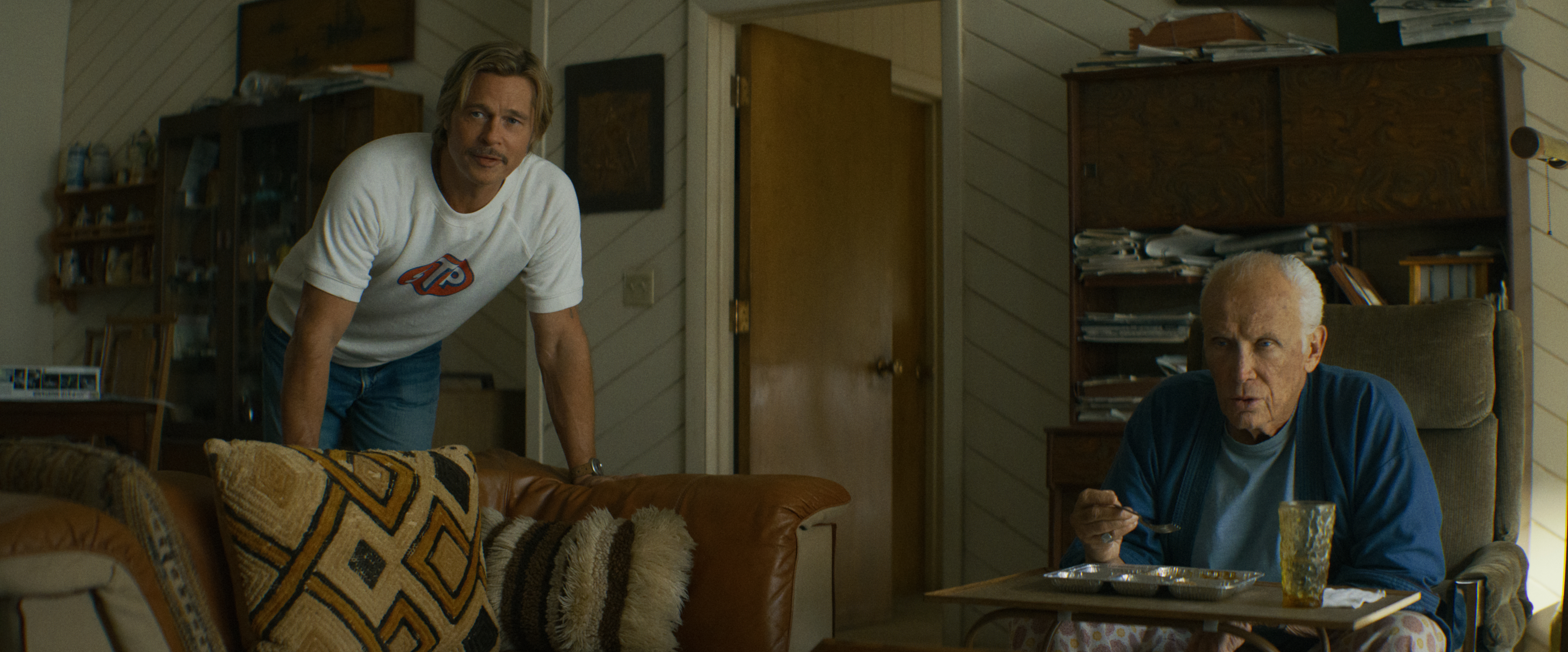 "Two men in a cozy living room, one sitting and eating at a tray table, the other leaning on a couch. The setting is warm with patterned pillows, wooden furniture, and shelves."