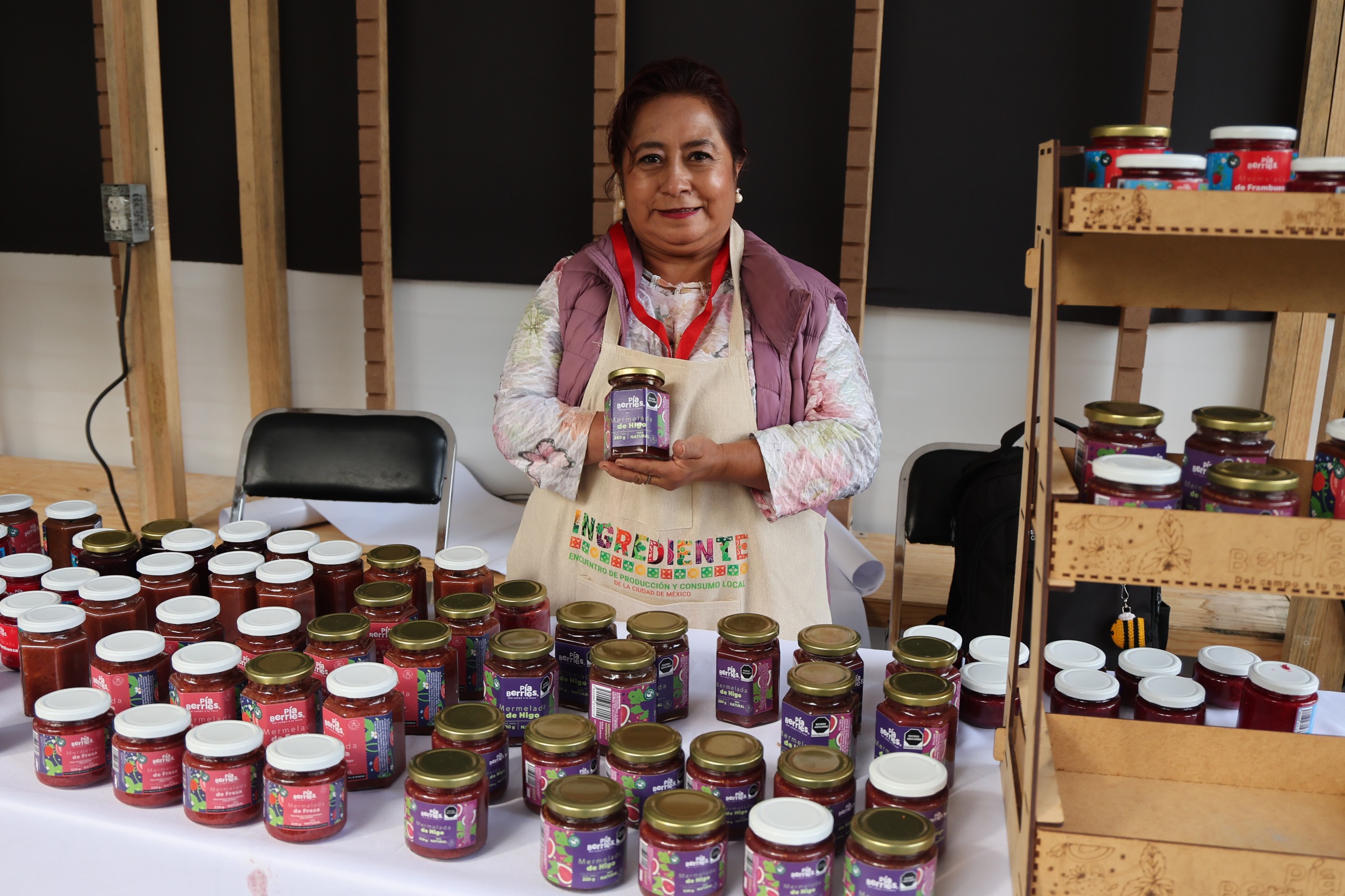 : Una mujer de mediana edad sonriente se encuentra detrás de un puesto de mercado bien surtido lleno de mermeladas artesanales. Sostiene un frasco de mermelada "Pío Berries" en sus manos y mira amigablemente a la cámara.