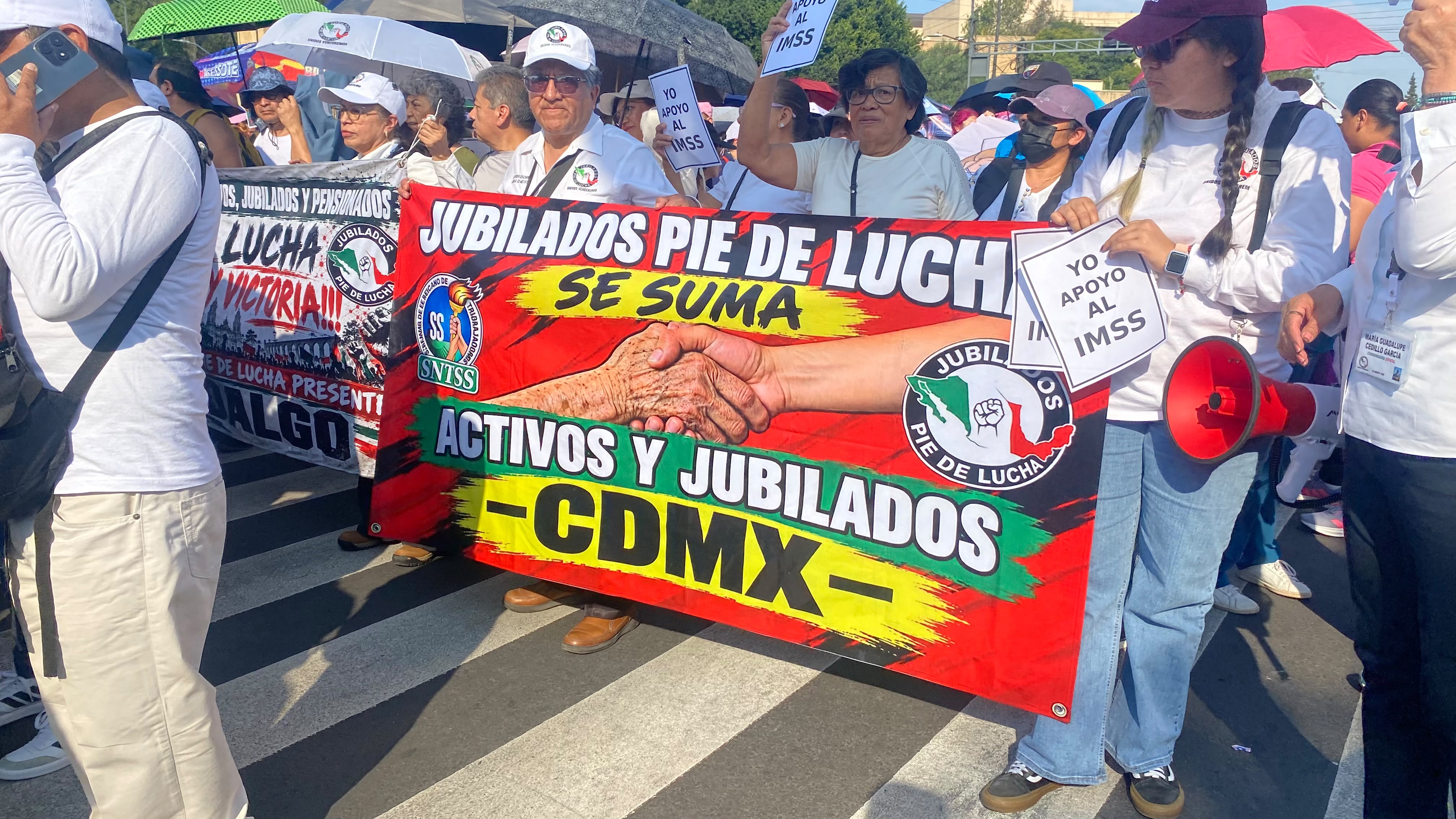 Texto Alternativo (Alt Text) para Accesibilidad y SEO: Una multitud de jubilados y trabajadores del IMSS manifestándose en una calle de la Ciudad de México. Sostienen una pancarta principal que dice "JUBILADOS PIE DE LUCHA SE SUMA ACTIVOS Y JUBILADOS -CDMX-". Otros manifestantes sostienen carteles de mano que dicen "YO APOYO AL IMSS". Una mujer sostiene un megáfono rojo. Es un día soleado.