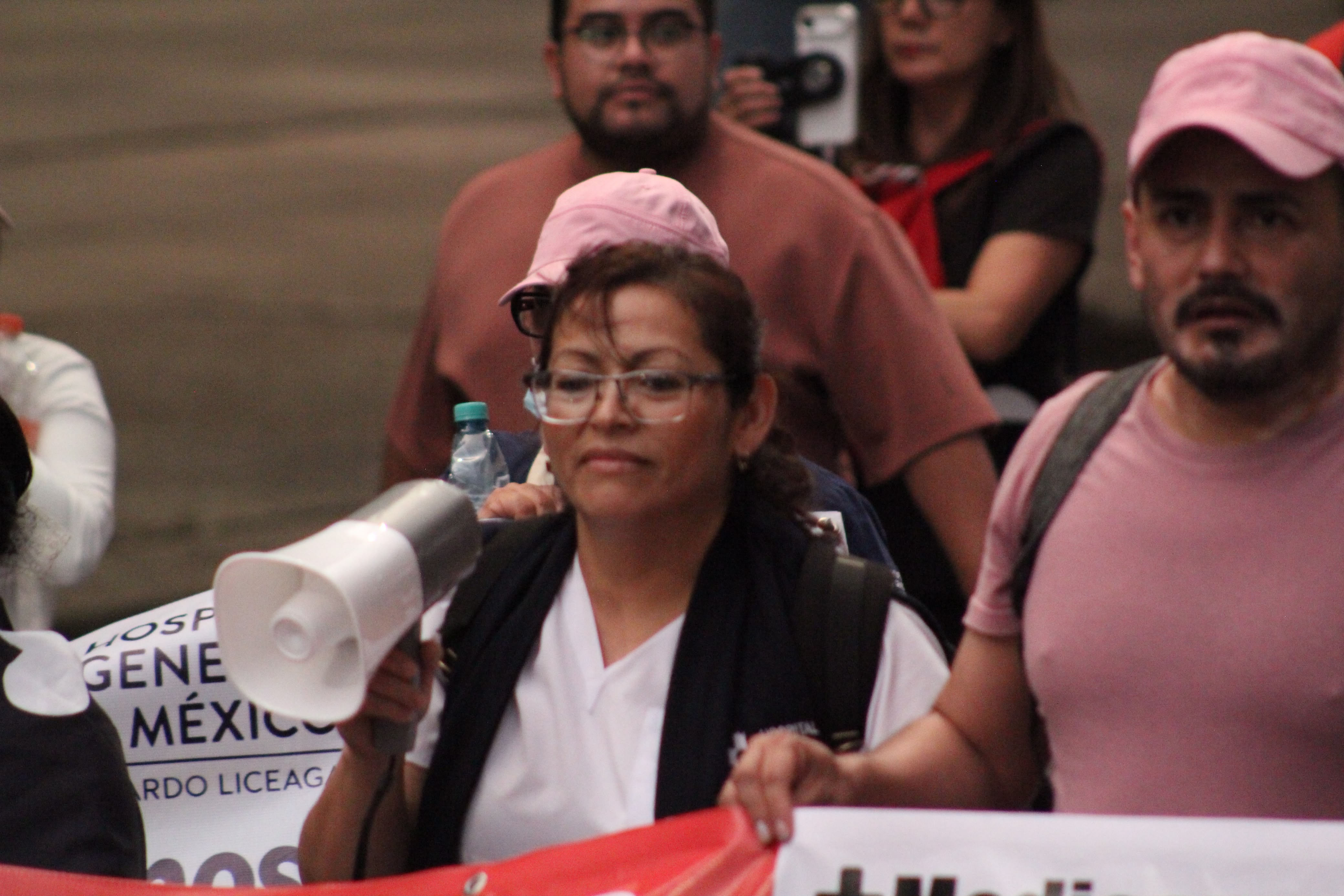 Una trabajadora de la salud con uniforme blanco y lentes sostiene un megáfono durante una marcha. Camina junto a otros manifestantes que llevan una pancarta del Hospital General de México.