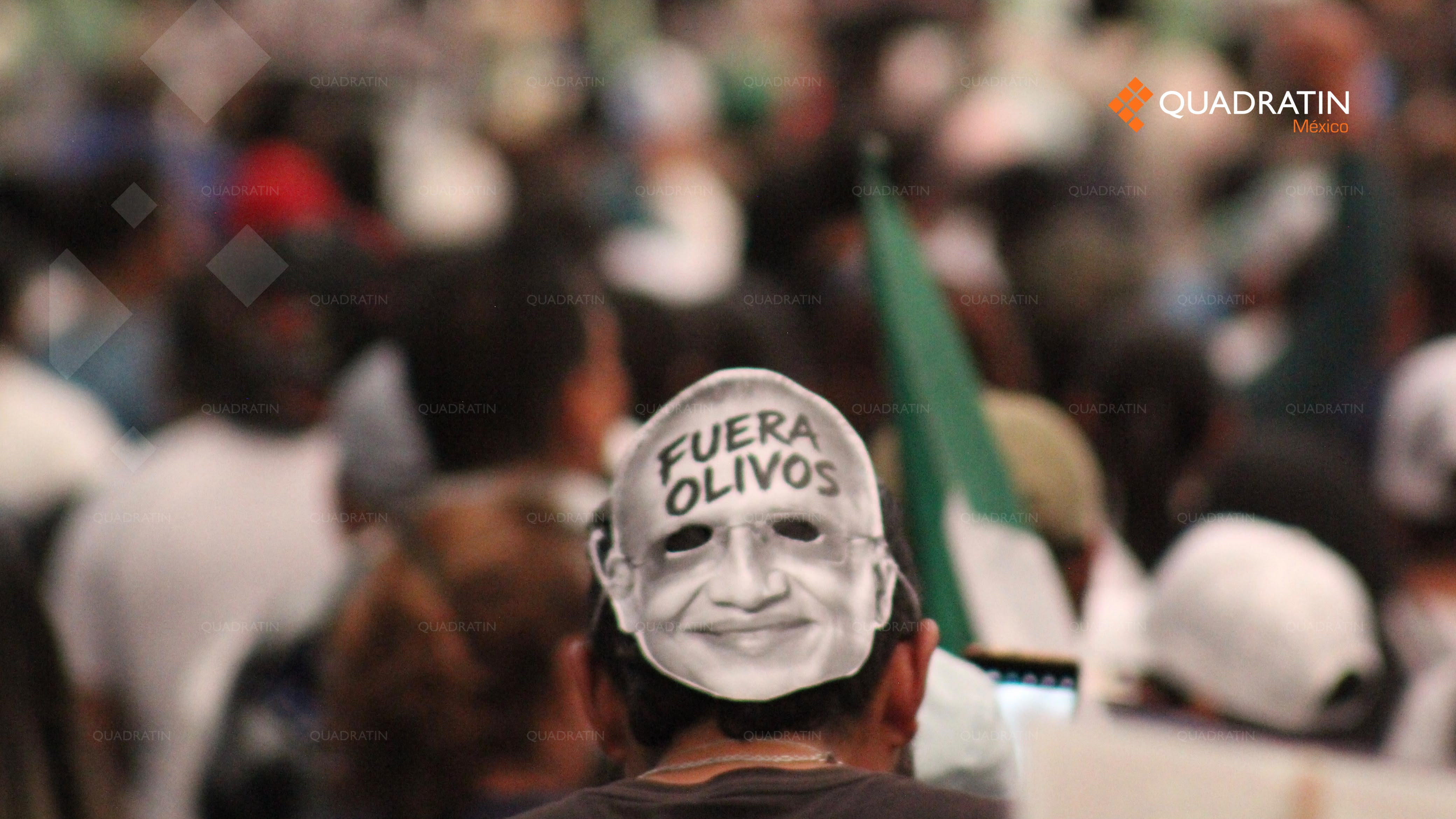 ersona de espaldas entre la multitud llevando una máscara impreso con el rostro de un hombre y la frase "FUERA OLIVOS" escrita en la frente, durante una protesta sindical del SNTSS.