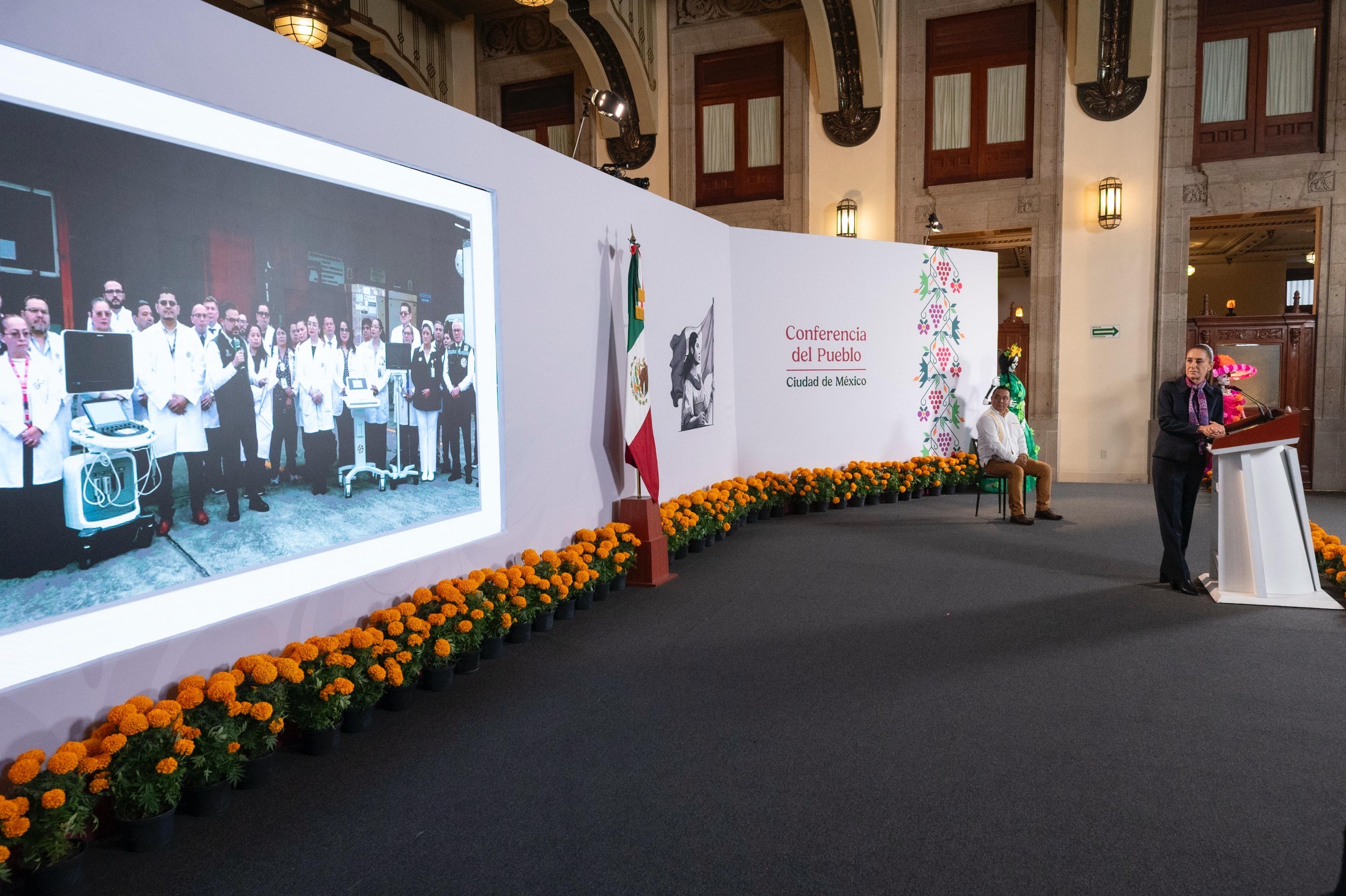 Claudia Sheinbaum en el pódium de la Conferencia del Pueblo junto a una pantalla con personal de salud y decoración de cempasúchil